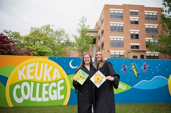 Sydnie Brown and Sarah Tower in graduation gowns in front of mural in their graduation robes